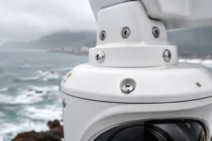Vue rapprochée d'une caméra de sécurité blanche avec des vis et un objectif, positionnée près d'une côte océanique sous un ciel couvert.